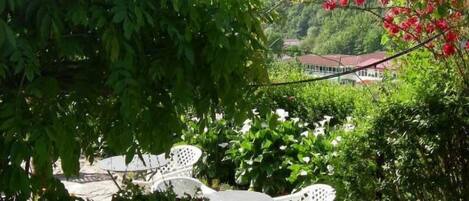 A garden patio with white plastic chairs and a table, surrounded by lush greenery and a small water feature.