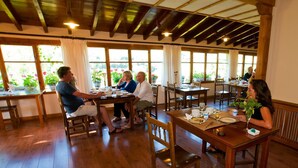 Un comedor con mesas y sillas de madera, ventanas amplias con cortinas y un techo rústico.