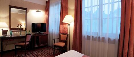 A hotel room with a bed, a desk with a computer, a chair, a TV, and a window with curtains.