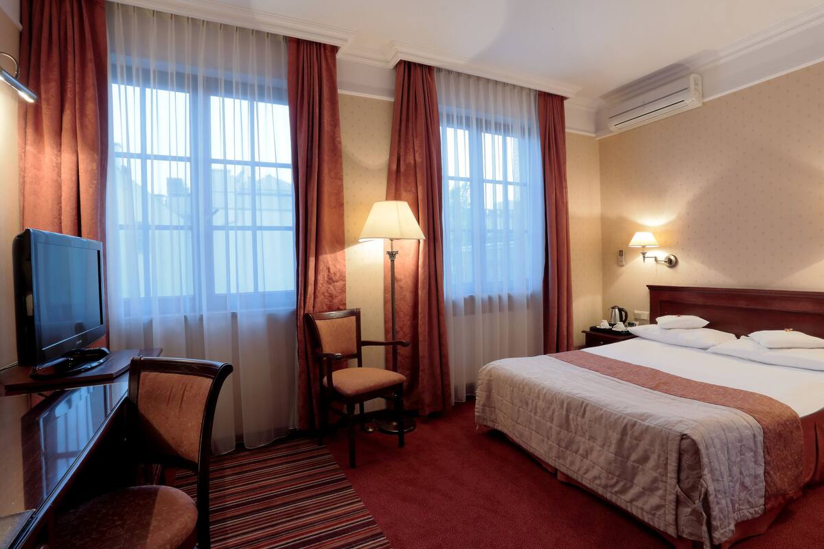 Ein Hotelzimmer mit einem Bett, einem Schreibtisch mit Fernseher, einem Sessel, einer Lampe und einem Nachttisch ebenfalls mit einer Lampe.