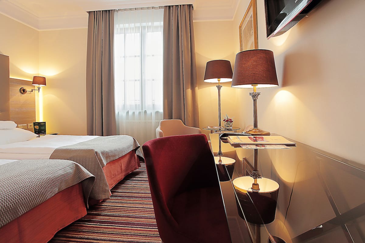Ein Hotelzimmer mit zwei Betten, einem Schreibtisch mit Lampe, einem Sessel und einem an der Wand montierten Fernseher.