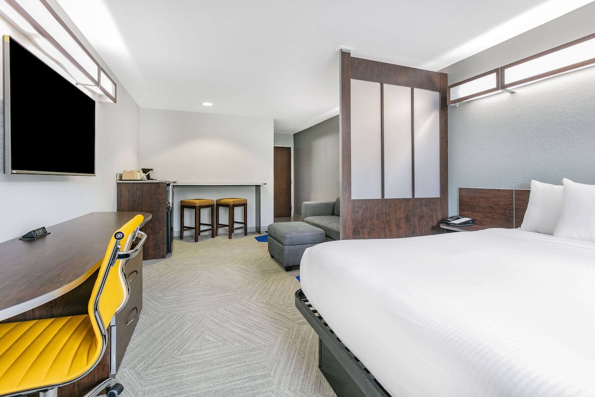 A hotel room with a bed, a desk with a chair, a TV, and a small seating area with stools.
