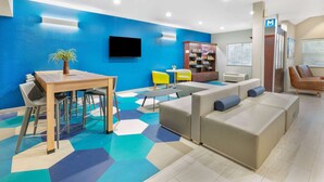 A modern hotel room with a blue accent wall, a flat-screen TV, a dining area with a table and chairs, a sofa, and a bookshelf. - Microtel Inn & Suites by Wyndham Austin Airport (Austin)