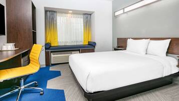 A hotel room with a bed, a desk with a chair, a window with curtains, and a TV mounted on the wall.