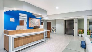 A reception area with a blue accent wall, a wooden reception desk, a seating area with blue cushions, and a bulletin board with framed pictures. - Microtel Inn & Suites by Wyndham Austin Airport (Austin)