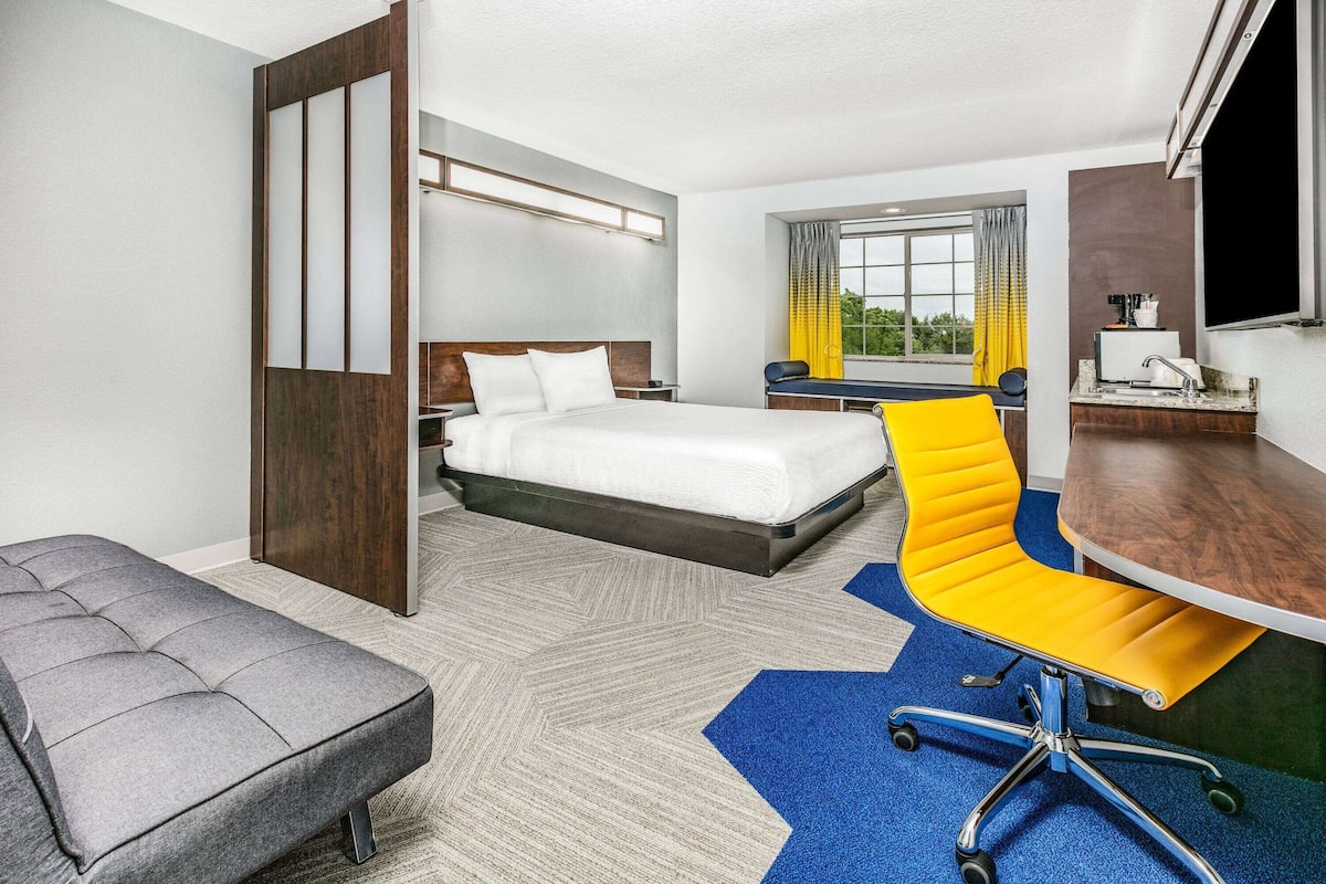 A hotel room with a bed, a desk, a chair, a bench, and a window with curtains.