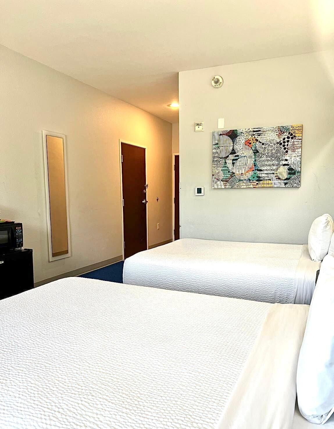 A hotel room with two beds, a door, a wall-mounted artwork, and a small table with a coffee maker.