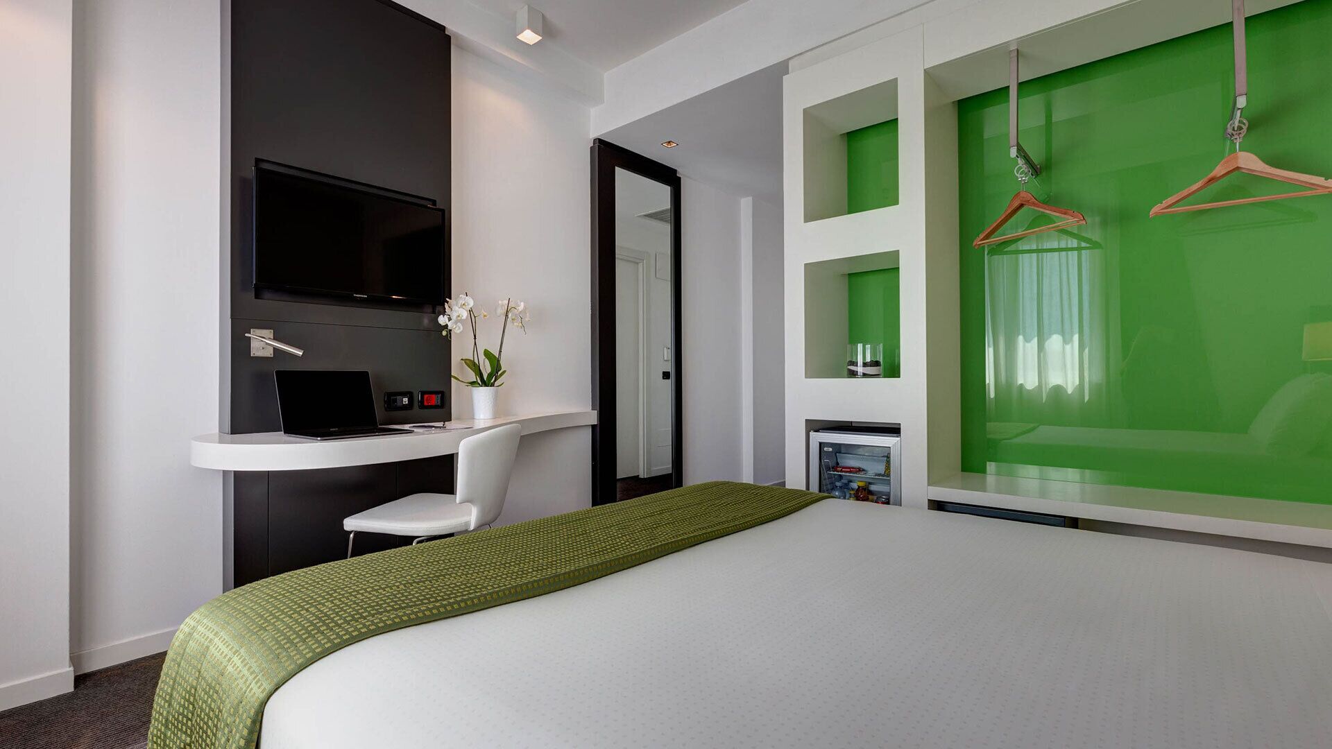 Une chambre d’hôtel moderne équipée d’un lit, d’un bureau avec un ordinateur portable, d’une télévision et d’un mur d’accent vert.