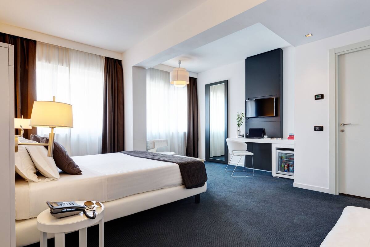 A hotel room with a bed, bedside table with a lamp, a desk with a computer, a minibar, and a door.
