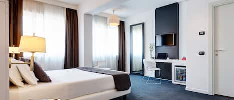 Habitación de hotel con cama, mesita de noche con lámpara, escritorio con computadora, minibar y puerta.