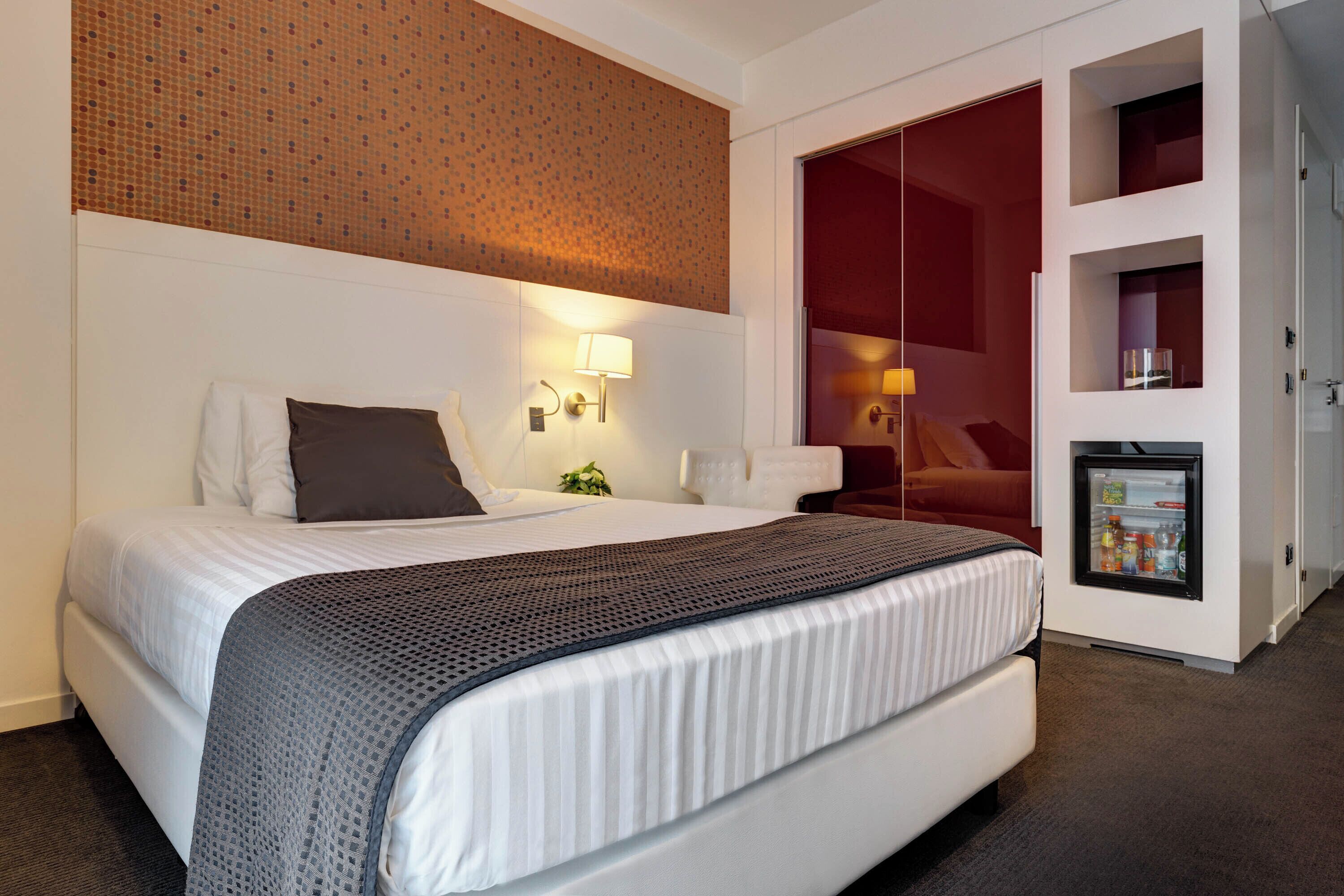 Une chambre d’hôtel avec un lit, une lampe fixée au mur, un petit réfrigérateur et une porte rouge.