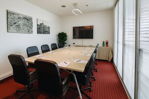Una sala conferenze con un lungo tavolo, sedie nere, una pianta e una TV appesa al muro.