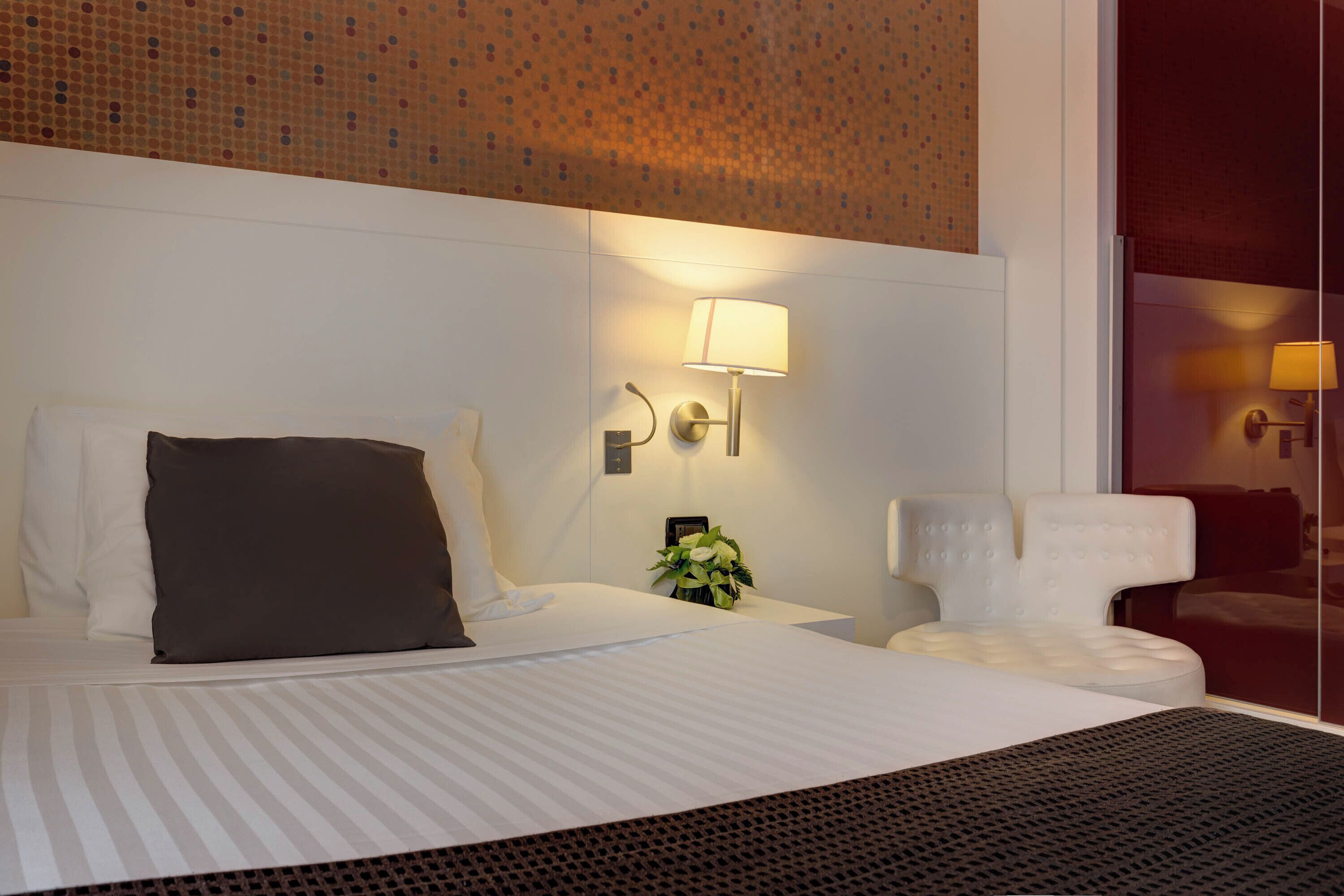A hotel room with a large bed, a bedside table with a lamp, and a view of the room's interior through a glass door.
