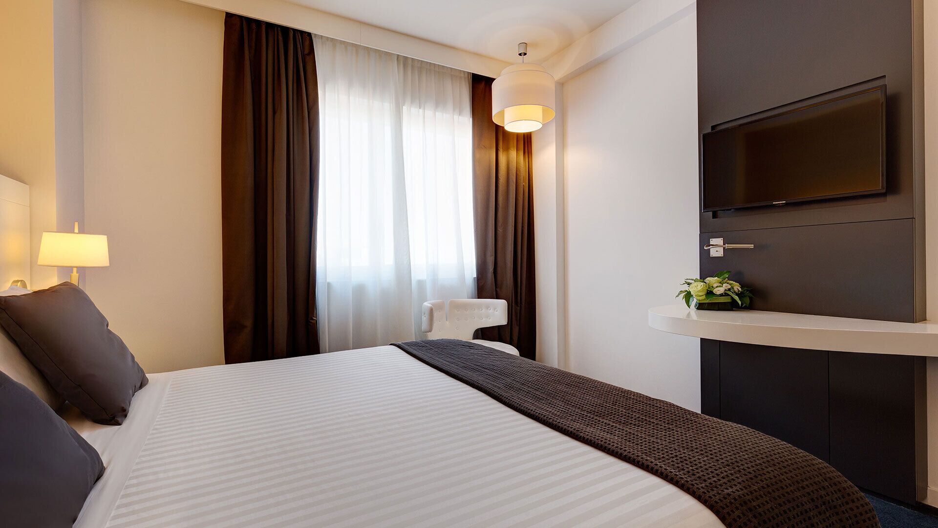 Habitación de hotel con una cama grande, un televisor de pantalla plana en la pared, una mesita de noche con lámpara y una ventana con cortinas.