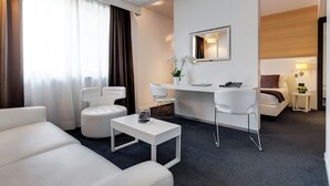A modern hotel room with a white sofa, a small white table, a desk with a chair, a bed, a mirror, and a nightstand with a lamp. - iH Hotels Roma Z3 (Rome)