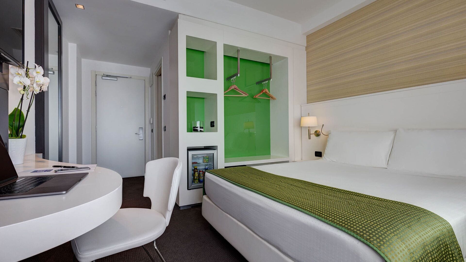 Ein Hotelzimmer mit Bett, Schreibtisch mit Laptop, Stuhl, Minibar und einer grünen Akzentwand.