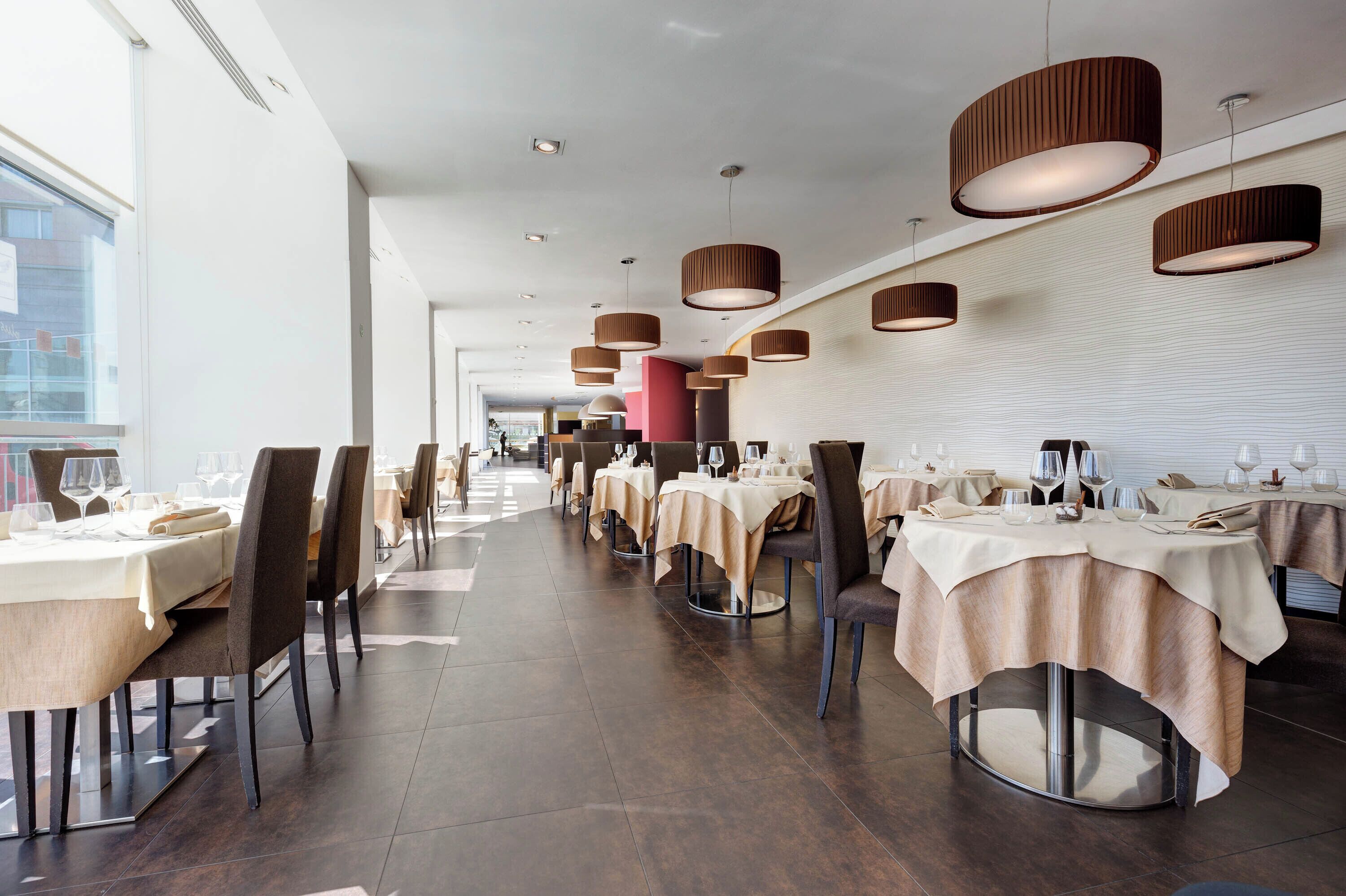 A modern restaurant with round tables, beige tablecloths, and brown pendant lights.