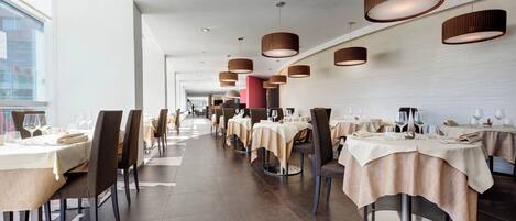 Un ristorante moderno con tavoli rotondi, tovaglie beige e lampade pendenti marroni.