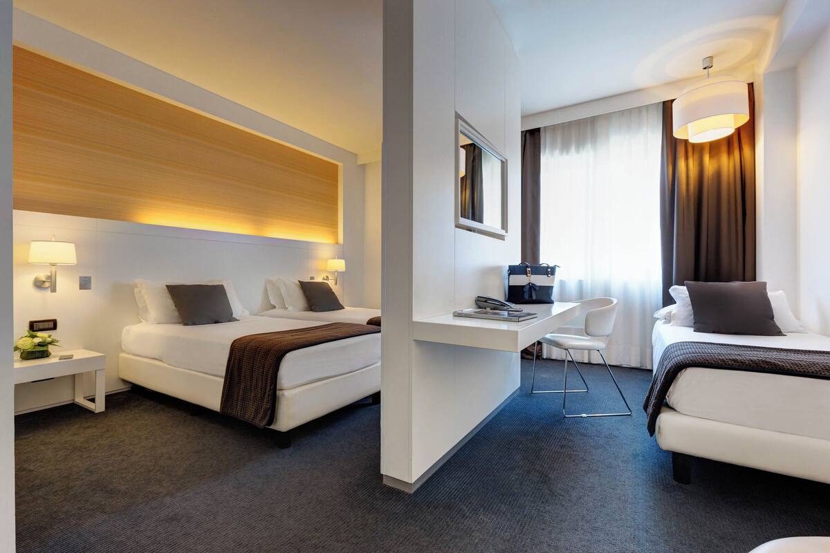 A hotel room with two beds, a desk, a chair, and a large window with curtains.