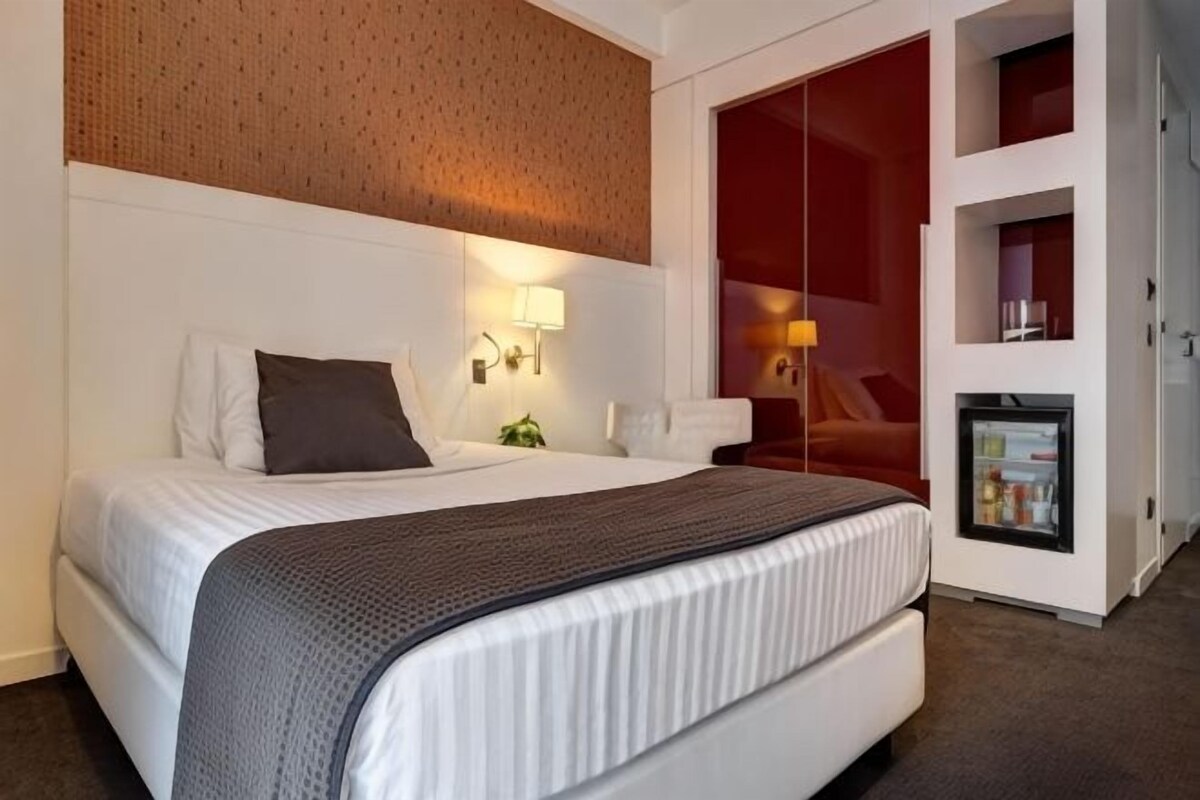 Ett hotellrum med en säng, ett sängbord, en lampa och ett litet kylskåp.