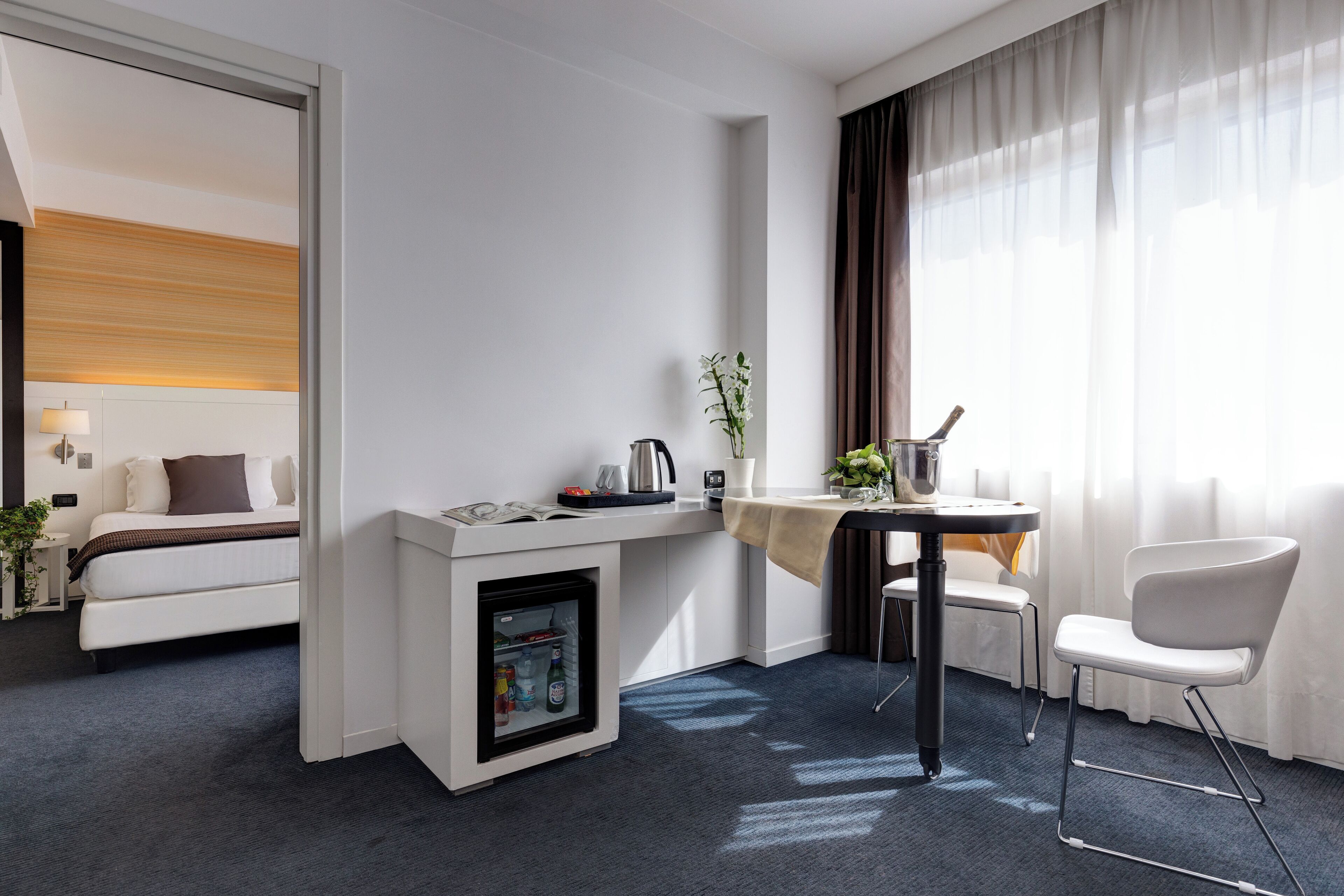 A modern hotel room with a bed, a mini-fridge, a table with a chair, and a window with curtains.