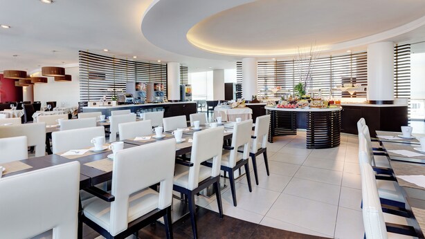 A modern hotel dining area with white chairs, tables, and a central island with a kitchen. - iH Hotels Roma Z3 (Rome)