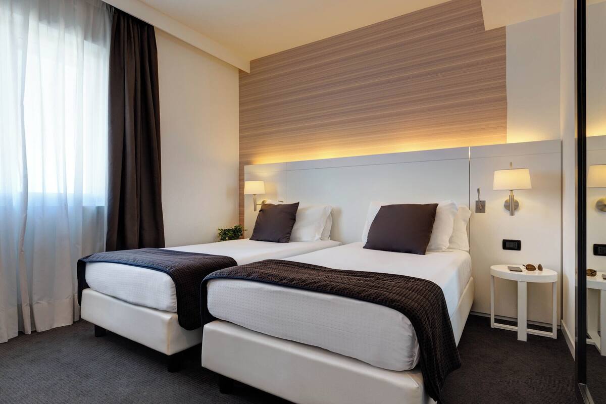 A hotel room with two beds, a small bedside table with a lamp, and a large mirror.