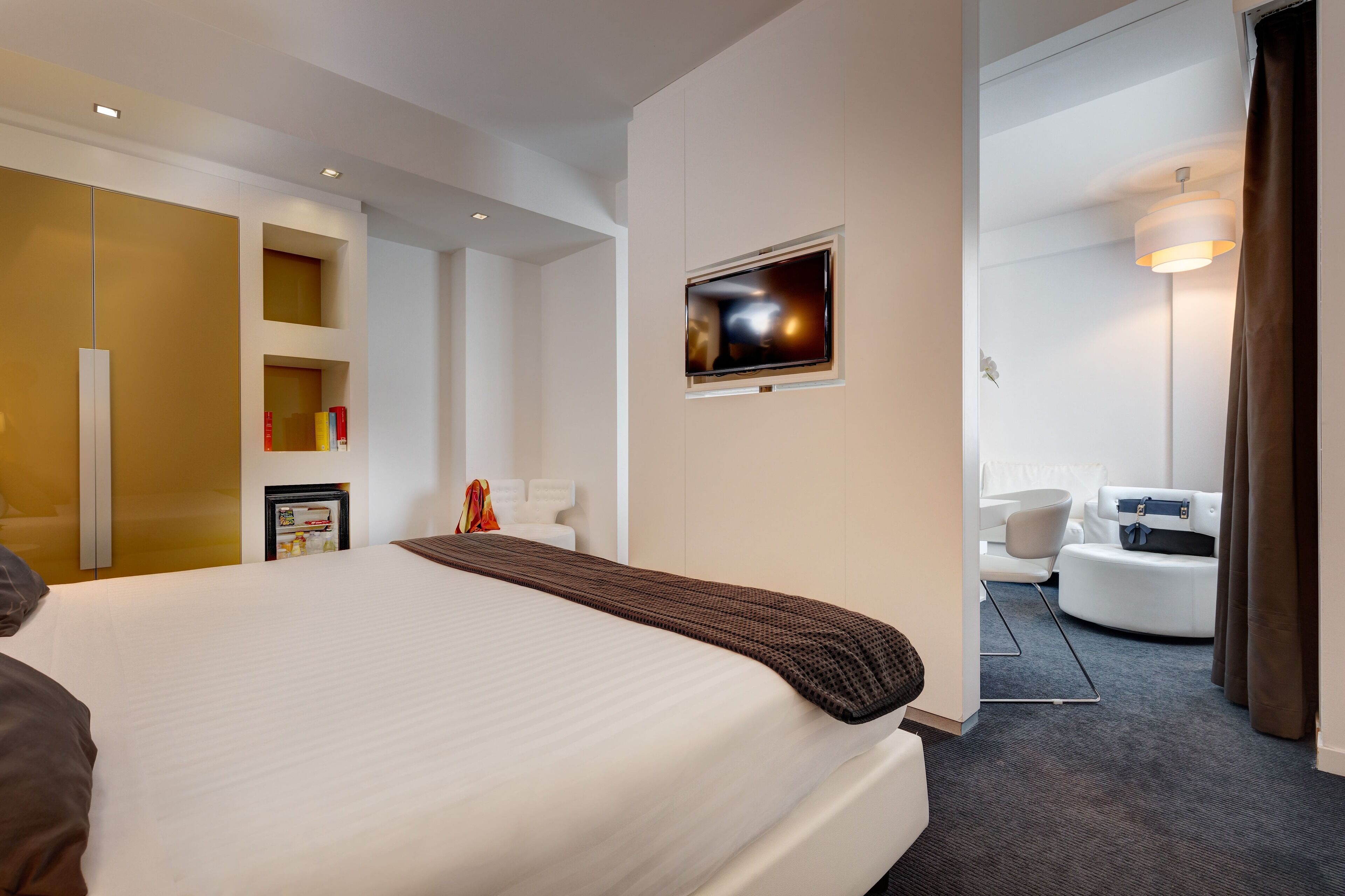 Quarto de hotel com uma cama grande, televisão presa na parede, um frigorífico pequeno e um espaço de estar com um sofá e poltrona.
