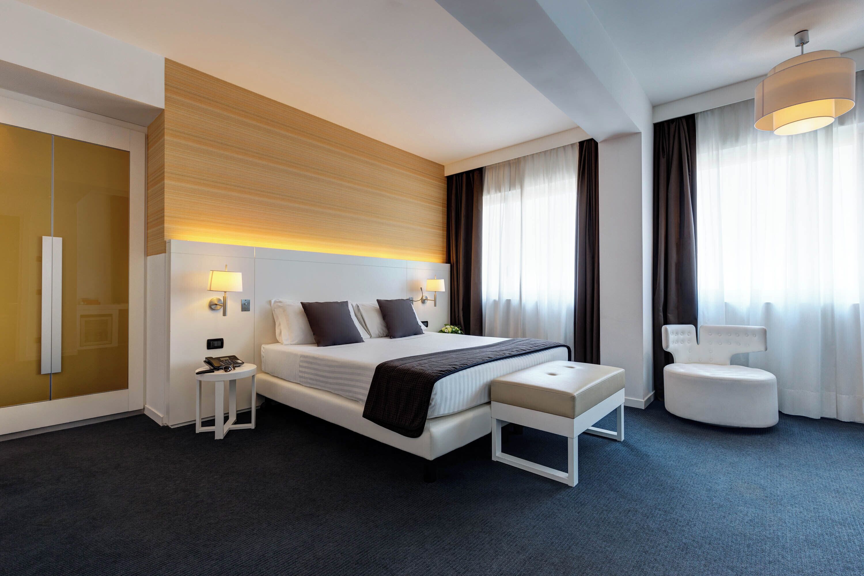 A modern hotel room with a large bed, bedside tables, a white bench, and a view of the city through large windows.