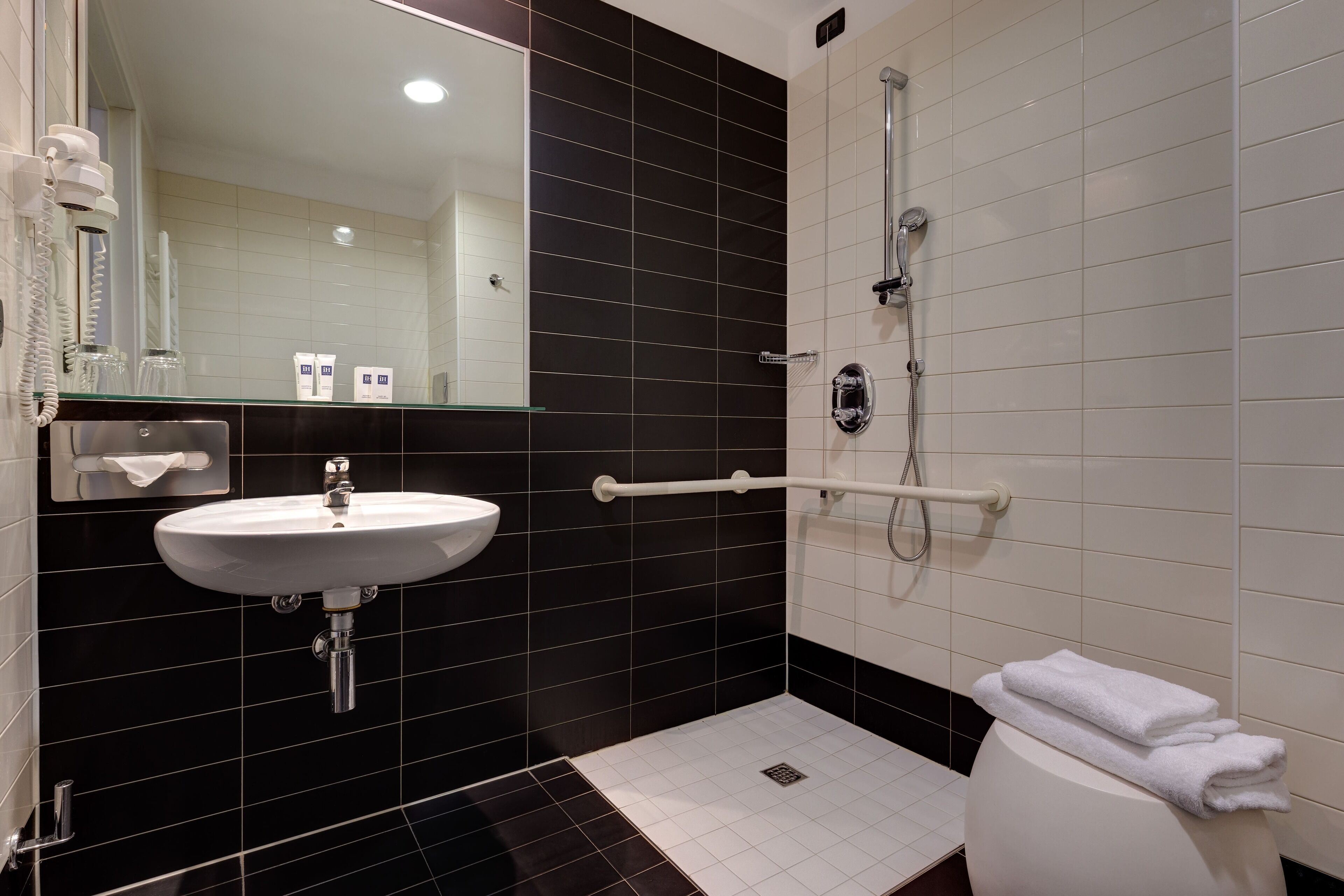 A modern bathroom with a white sink, black and white tiled walls, a glass shower enclosure, and a white towel on the toilet.