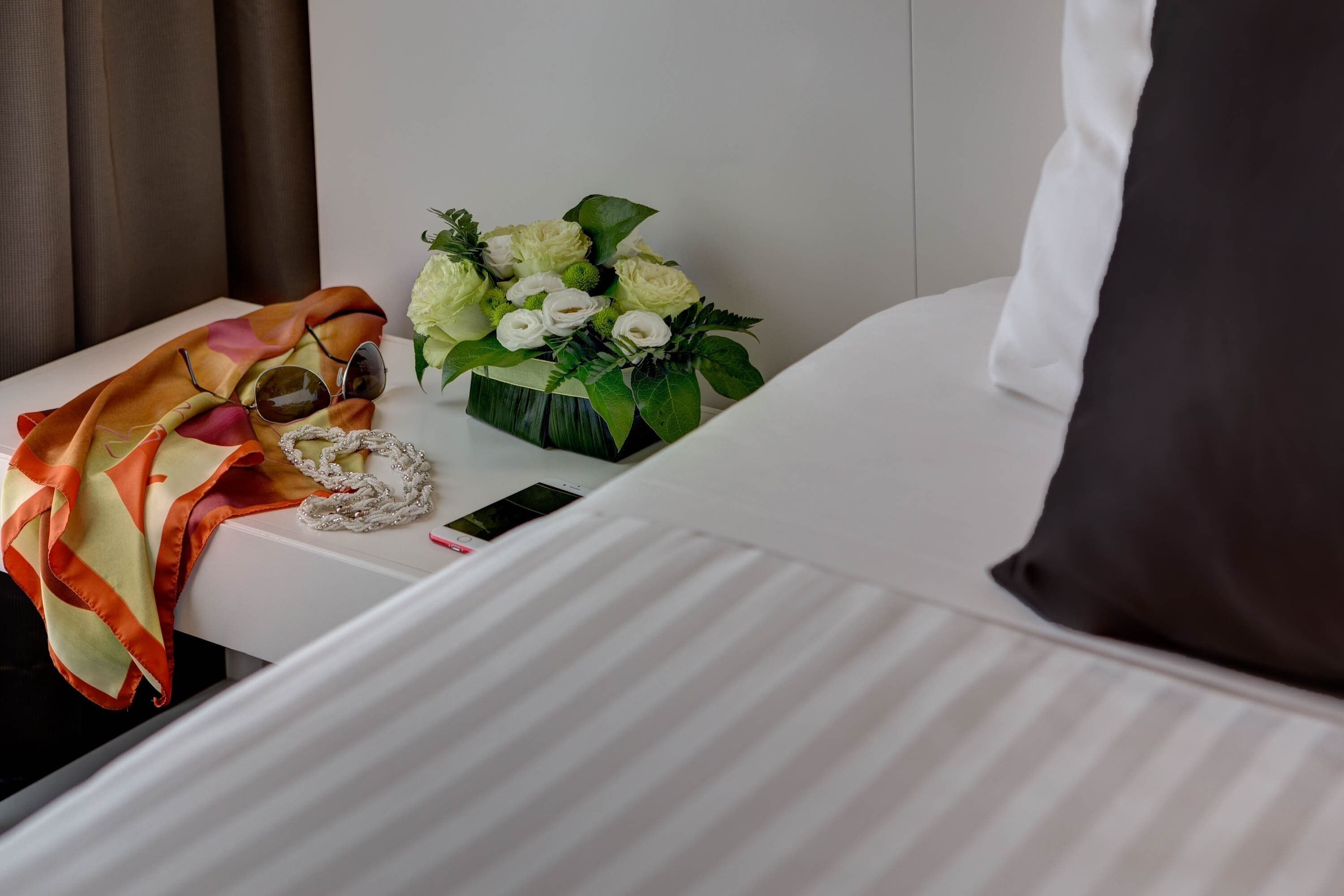Ein ordentlich eingerichtetes Hotelzimmer mit einem Bett, einem Nachttisch mit Blumenvase, einer Sonnenbrille und einem dekorativen Gegenstand.