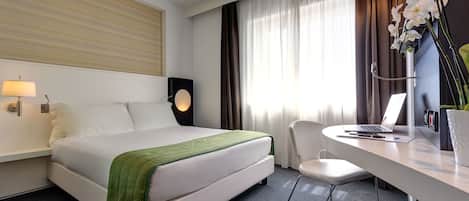 A hotel room with a bed, a desk with a laptop, a chair, a lamp, and a plant.