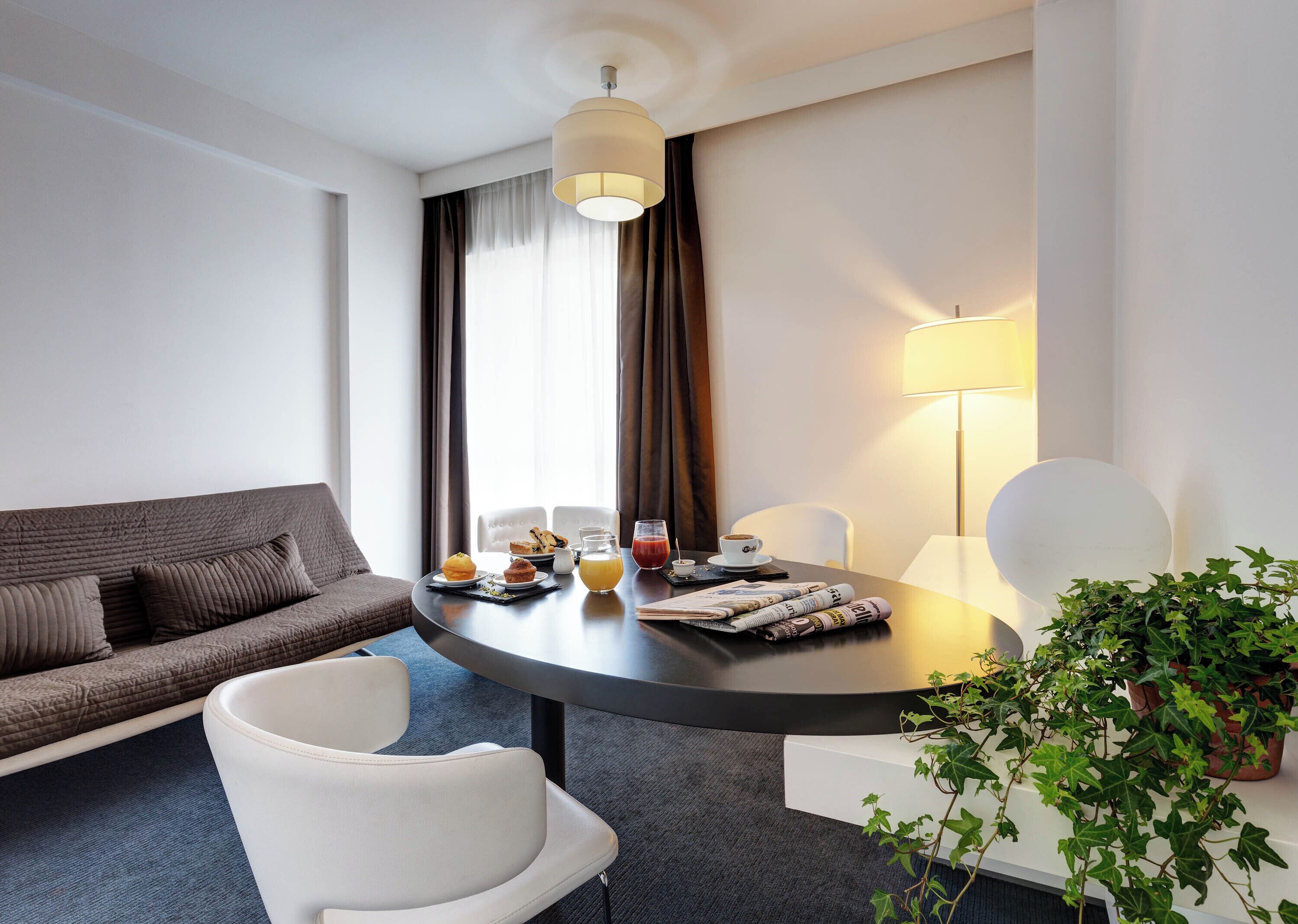 Una habitación de hotel moderna con una mesa de comedor redonda, un sofá y una planta en maceta.