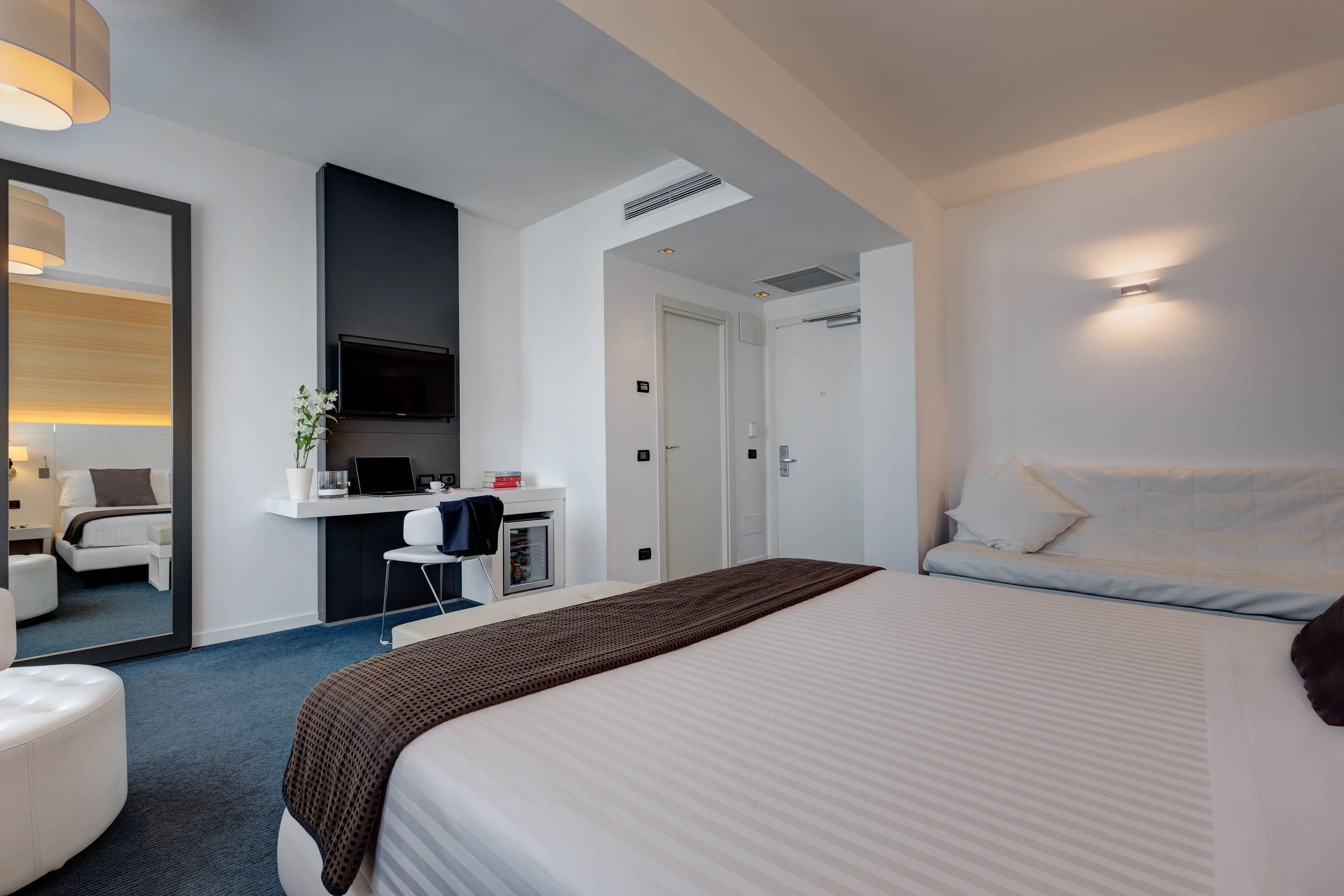 Una habitación de hotel moderna con una cama grande, un escritorio con ordenador, un televisor y una zona de estar separada con una silla y una mesita.