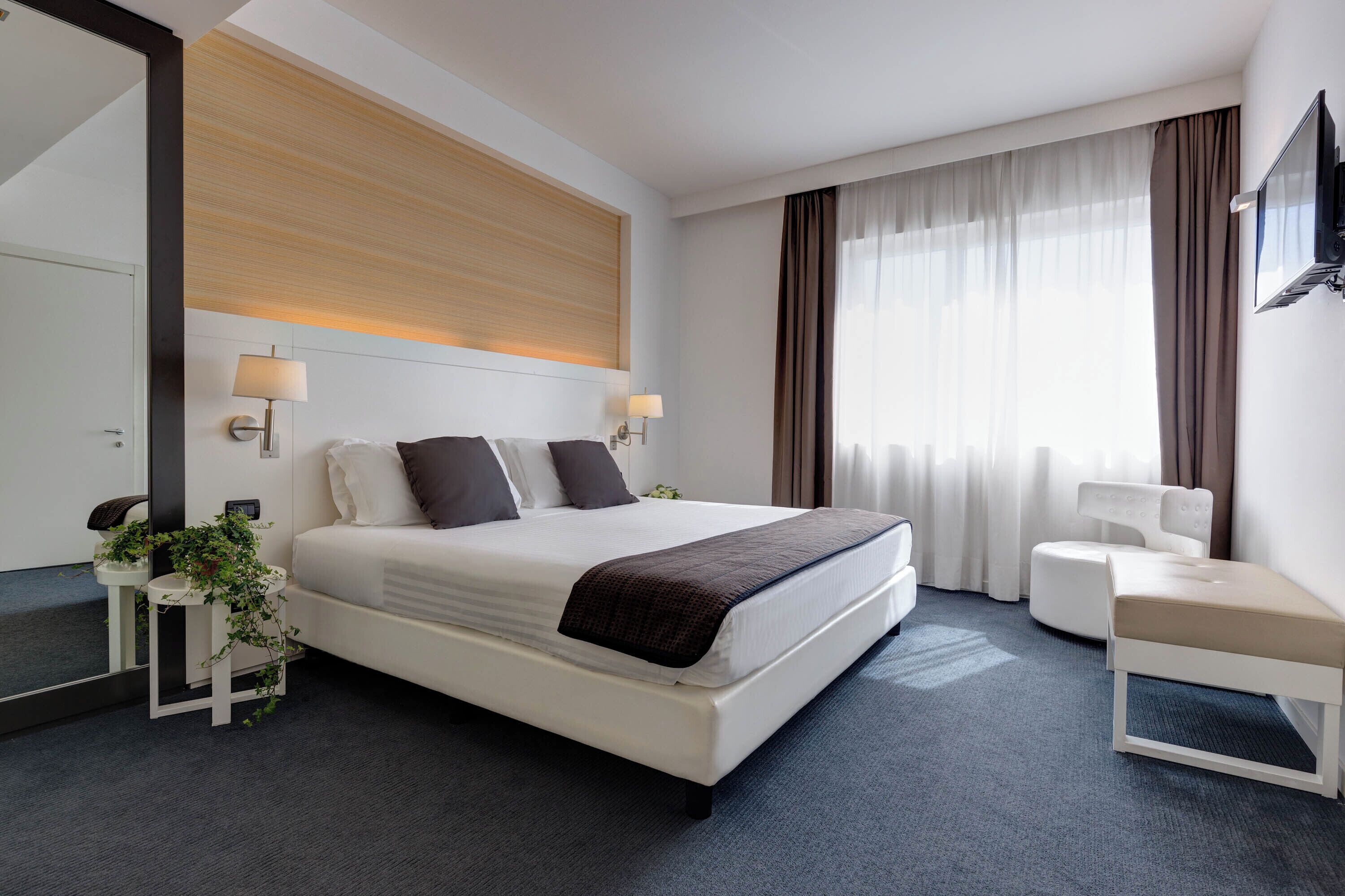 Una camera d'albergo moderna con un letto grande, lampade sui comodini, un tavolino con una pianta, una panca e un televisore fissato alla parete.