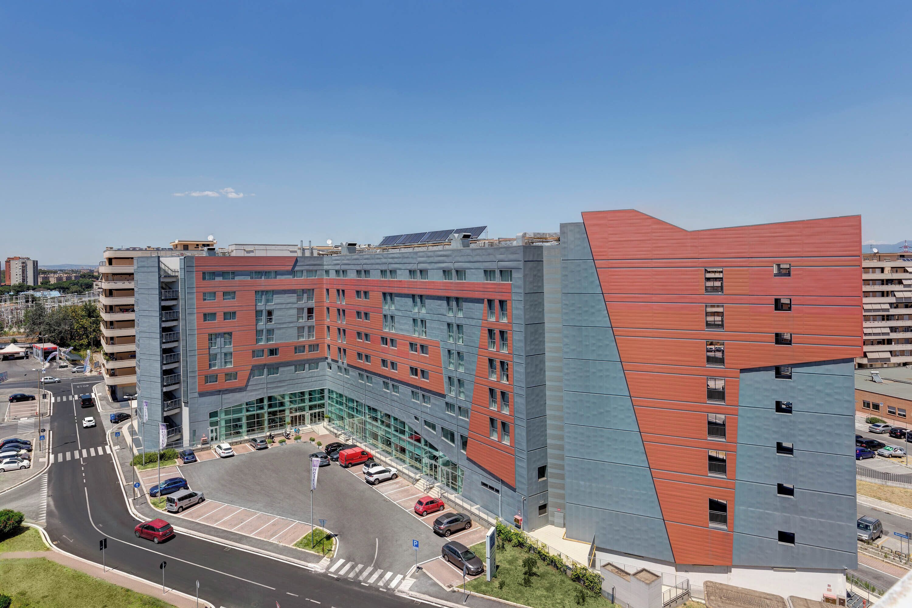 A modern building with a unique angular design, featuring multiple floors and a parking area with several cars.