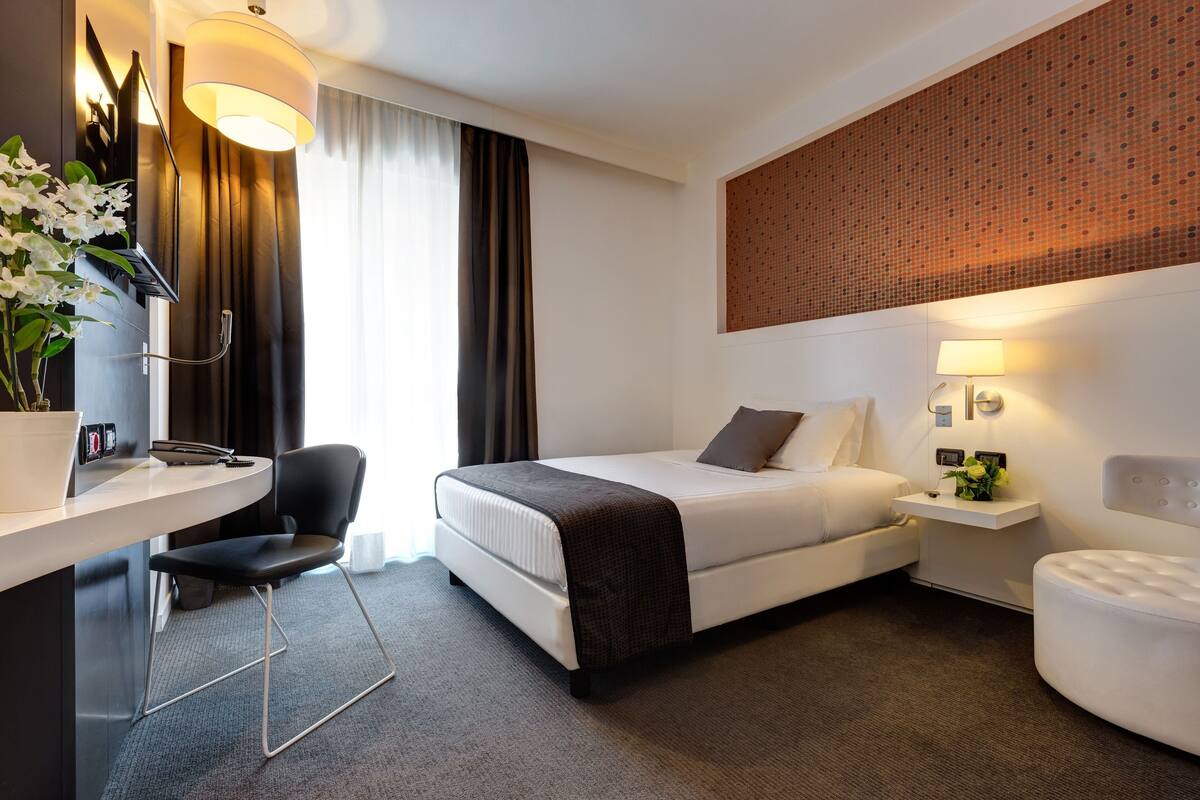A modern hotel room with a bed, a desk, a chair, a bedside table with a lamp, and a decorative wall panel.