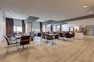 A spacious conference room with multiple tables and chairs, a kitchenette area, and a large window with curtains.