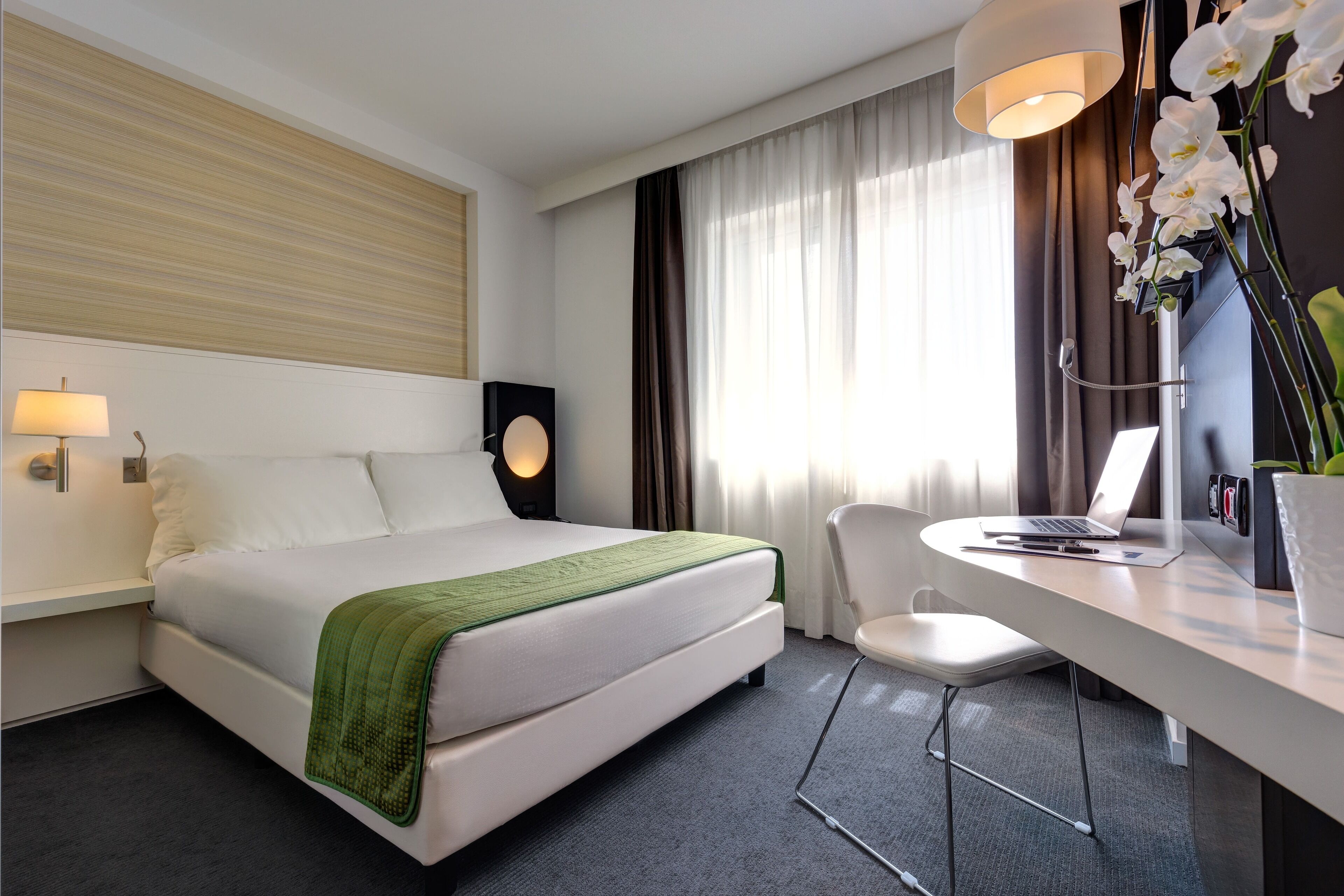 A modern hotel room with a neatly made bed, a desk with a laptop, and a chair.