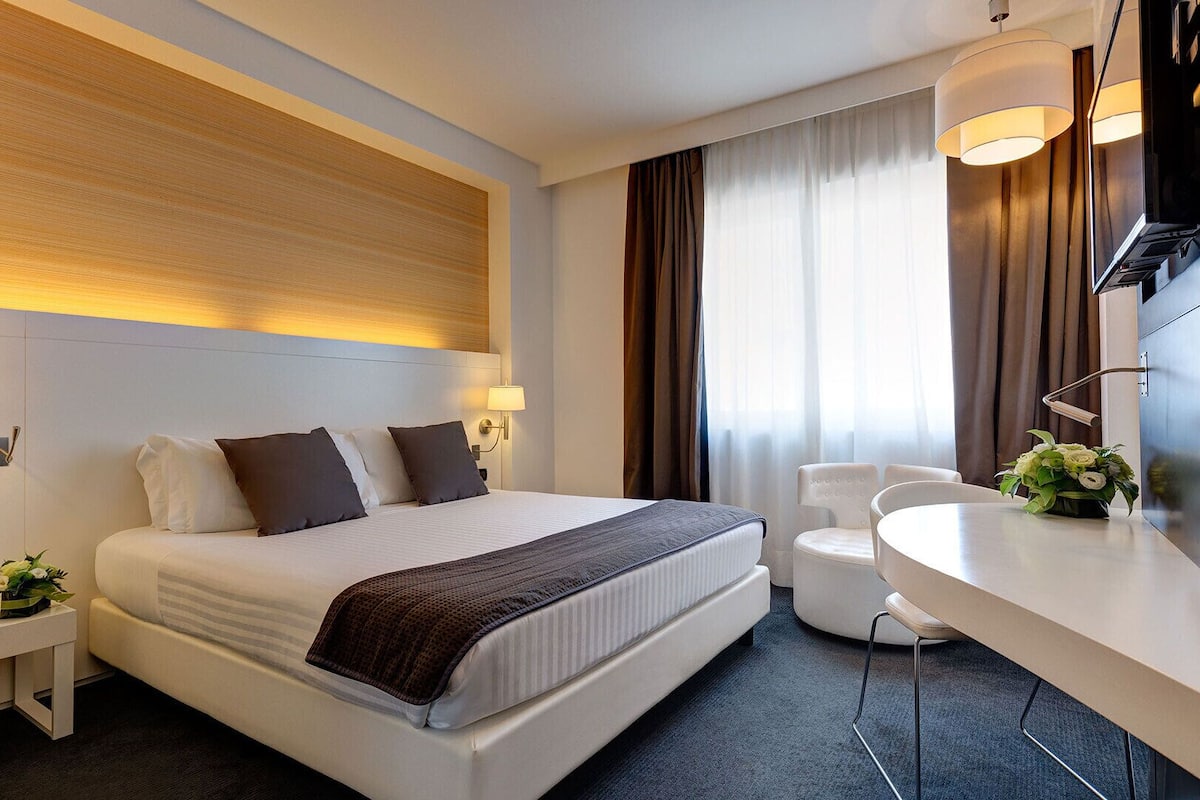 A modern hotel room with a large bed, bedside lamps, a desk with a chair, and a large window with curtains.