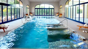 Una piscina interior con agua cristalina, iluminada por amplios ventanales, rodeada de sillas de descanso y toallas.