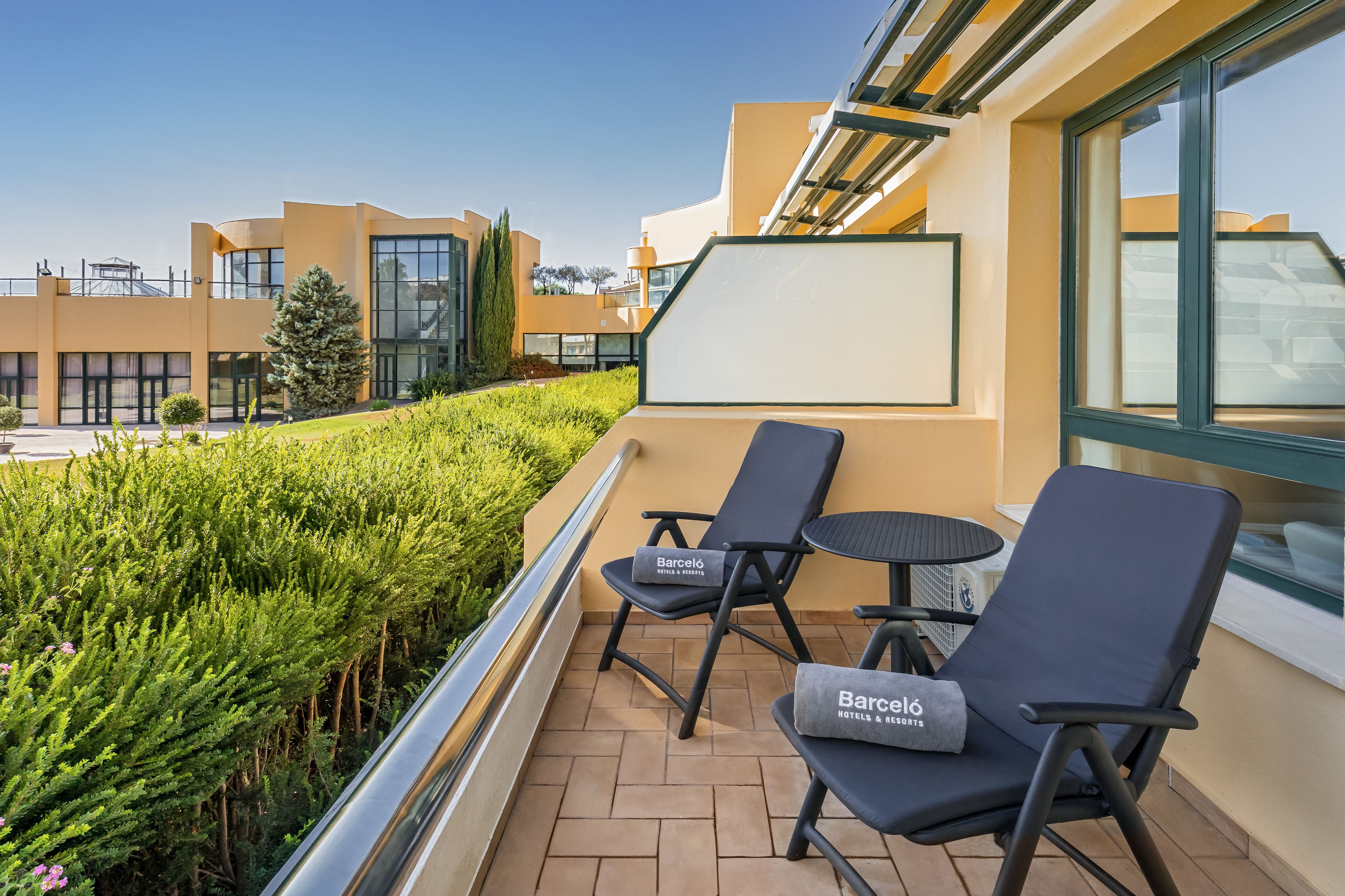 A balcony with two chairs and a small table, a clear view of the surrounding buildings, and a neatly trimmed hedge.