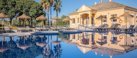 A pool with lounge chairs and umbrellas, a building with arches, and palm trees in the background.