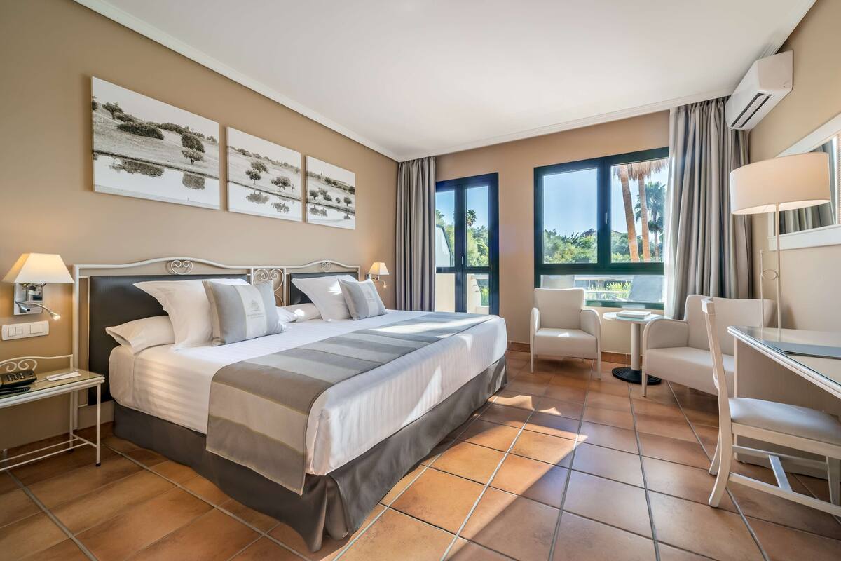 Ein Hotelzimmer mit einem großen Bett, zwei Nachttischen, einem Schreibtisch, einem Sessel und Blick ins Freie.