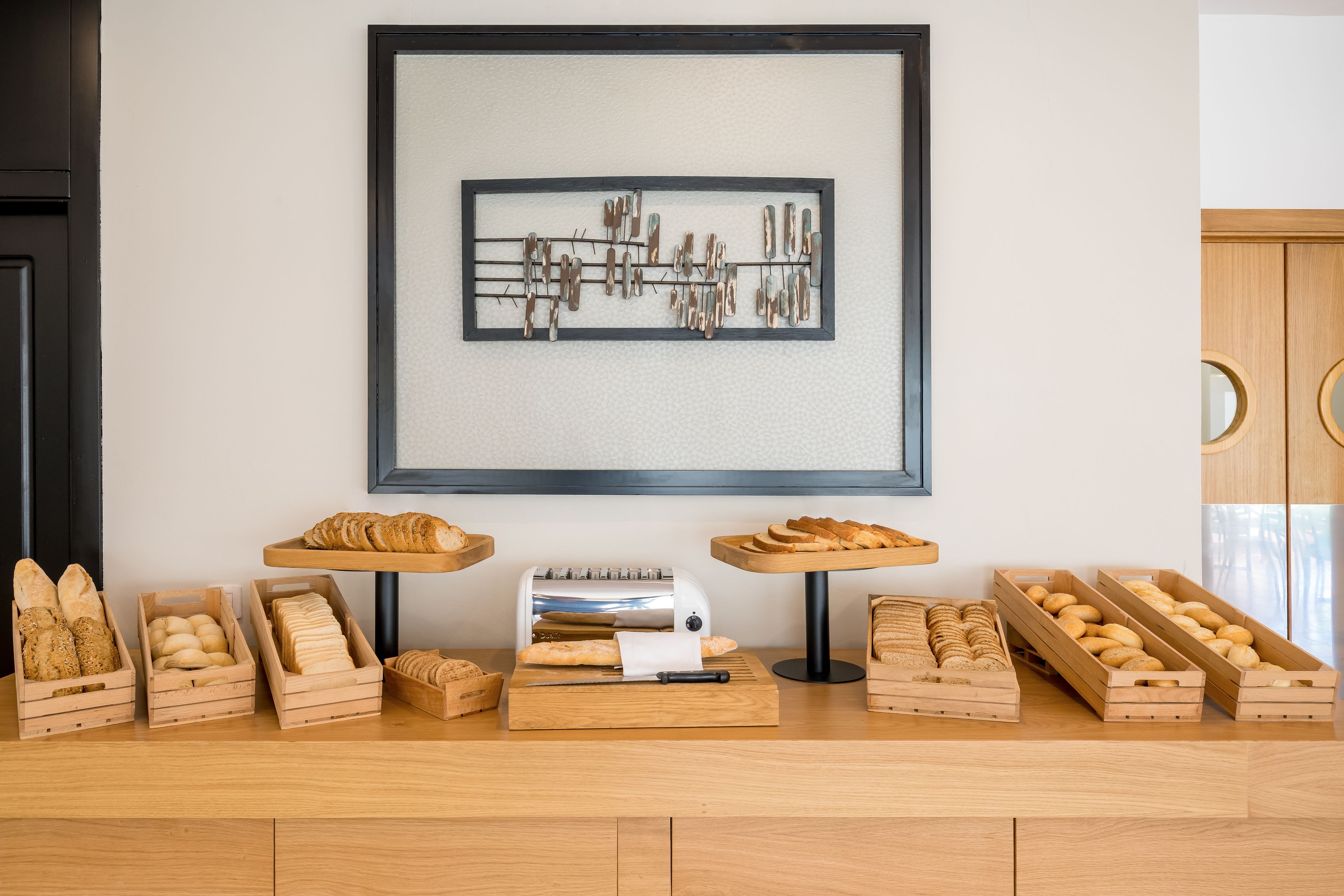 A bakery display with various breads and pastries in wooden crates, a bread slicer, and a framed artwork on the wall.