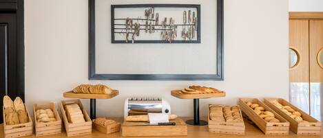 A bakery display with various breads and pastries in wooden crates, a bread slicer, and a framed artwork on the wall.