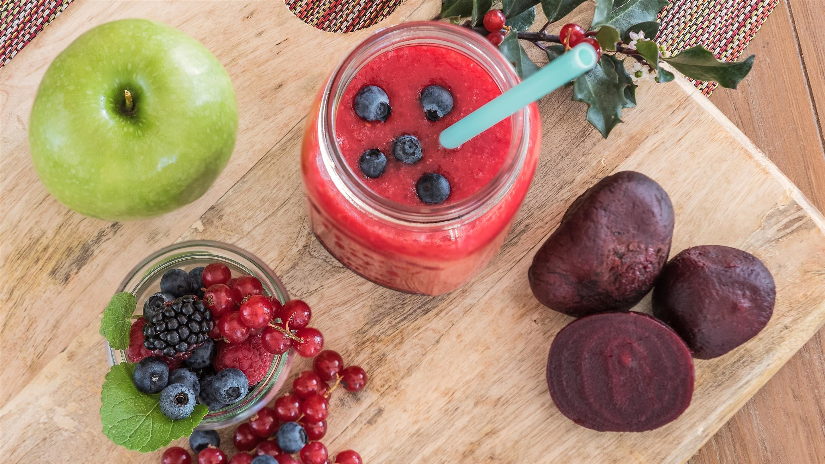 A wooden table with a green apple, a glass jar with a red smoothie and blueberries, a bowl of mixed berries, and sliced beets.