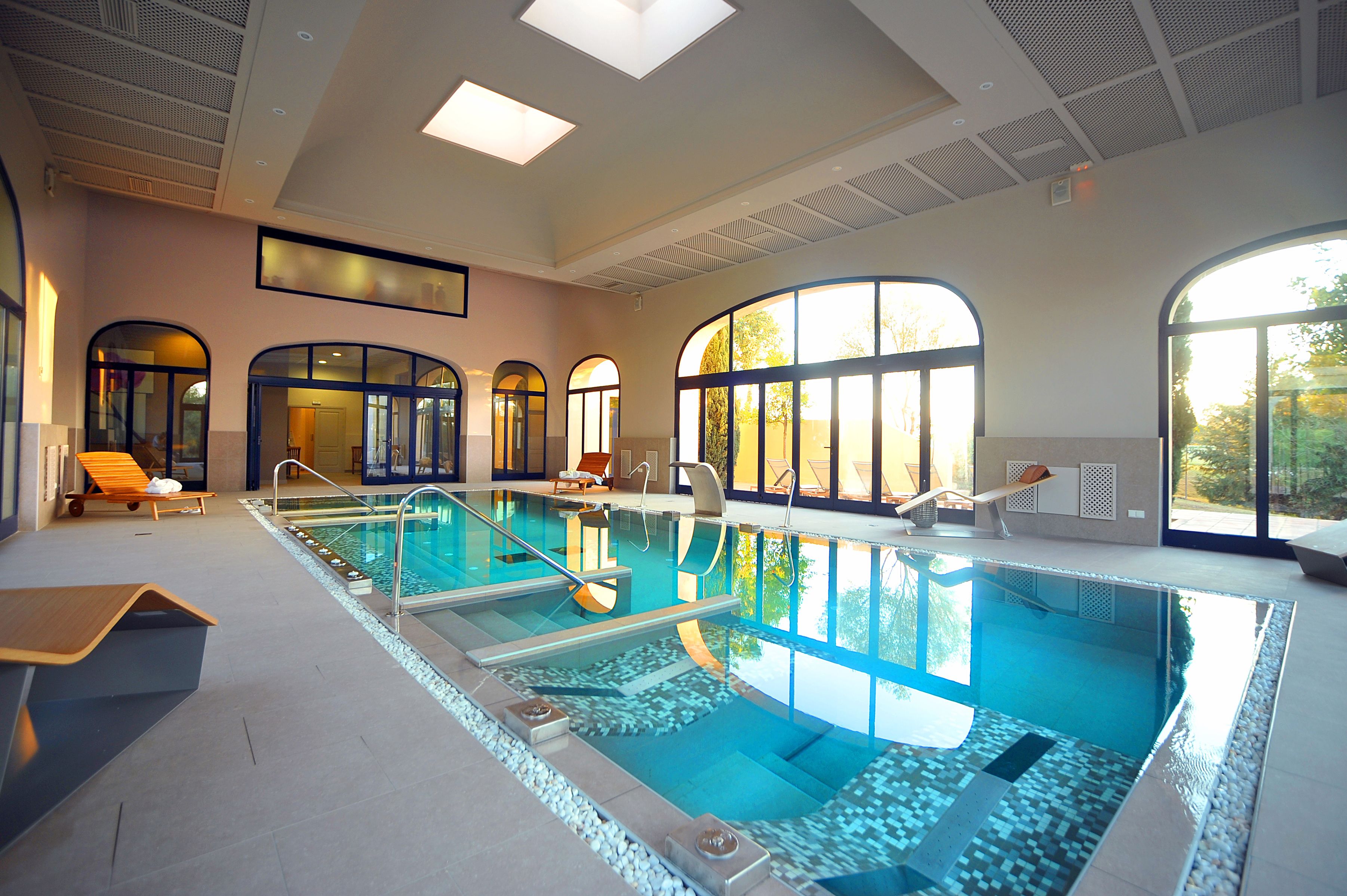 A large indoor swimming pool with clear water, surrounded by lounge chairs and a sunlit room with large windows.