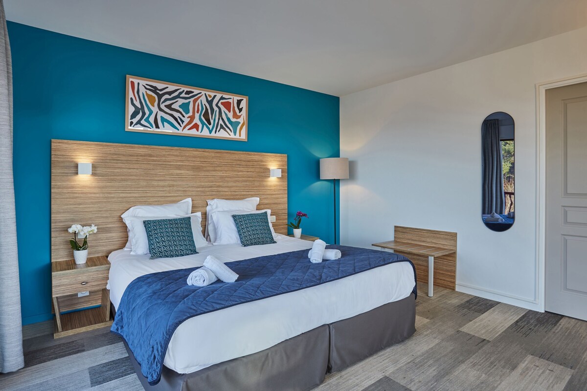 Un lit bien fait, avec une couette bleue, des oreillers blancs et une tête de lit en bois. Une petite table avec une lampe se trouve à côté du lit, et une fenêtre offre une vue sur la verdure.