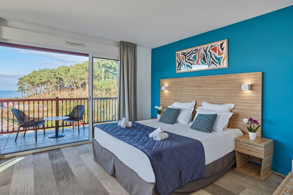 Une chambre avec un grand lit, une tête de lit en bois, un balcon avec des chaises et une vue sur la mer.